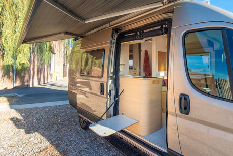 RV rental: Open side of a modern camper van with extended awning, revealing its interior kitchen and fold-out table.