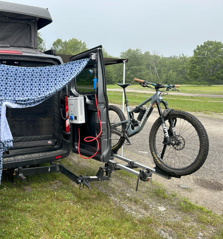 This RV rental camper van features a pop-top, outdoor shower, and a mountain bike on its rear rack, parked in a grassy area.