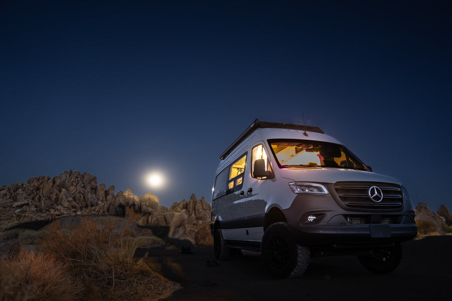 Moonlit desert camping with a bright Mercedes-Benz Sprinter RV rental under a starry sky. Interior lights glow invitingly.