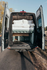 Open rear doors of a grey RV rental camper van reveal a comfy bed, overhead lighting, and storage.