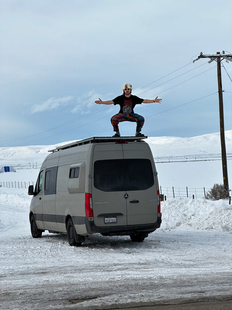Person in a fur hat squats on an RV rental Mercedes Sprinter in a snowy, hilly landscape, ready for a winter adventure.