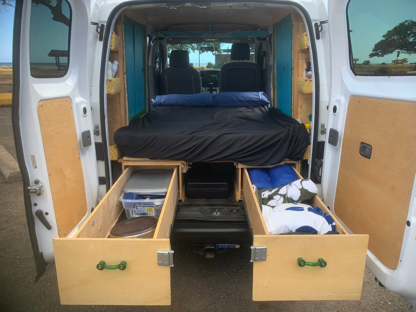Inside a custom RV rental: a compact camper van interior featuring a cozy bed and pull-out storage, parked by the beach.
