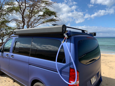 This blue RV rental van, parked on a sandy beach, includes a handy roof-mounted outdoor shower system.
