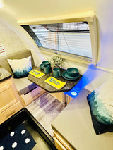 This compact RV rental features a cozy dinette with teal place settings, comfy seating, and ambient blue lighting.