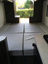 An RV rental interior with light gray cushions arranged as beds, looking out through the open rear doors to a sunny landscape.