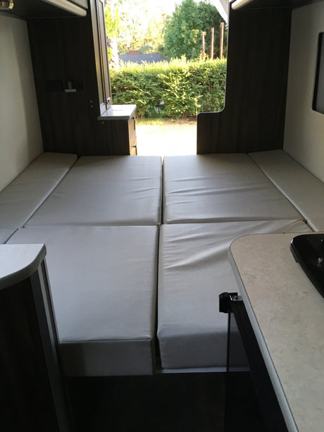 An RV rental interior with light gray cushions arranged as beds, looking out through the open rear doors to a sunny landscape.