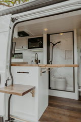 Two fold-out wooden tables extend from white cabinetry in this modern RV rental's kitchen and shower area.