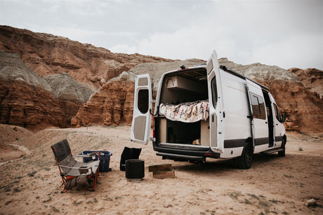White RV rental parked in a colorful desert landscape with open doors, ready for remote camping. Outdoor gear visible.