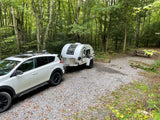 White SUV towing a teardrop RV rental at a wooded campsite with gravel, rocks, and picnic table.