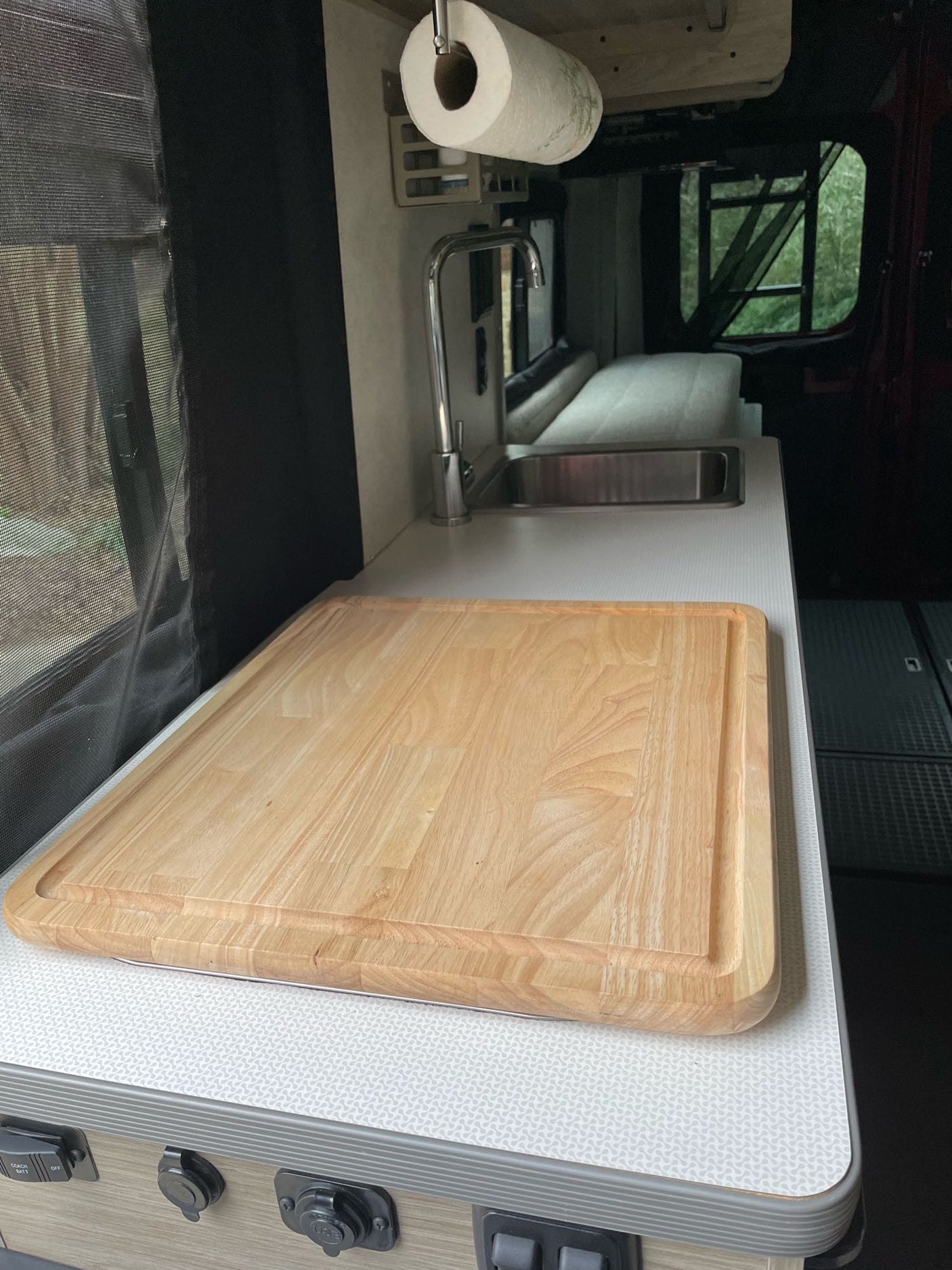 Inside an RV rental: a kitchen counter with a large wooden cutting board, sink, and screen window. Green nature outside.
