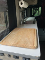 Inside an RV rental: a kitchen counter with a large wooden cutting board, sink, and screen window. Green nature outside.