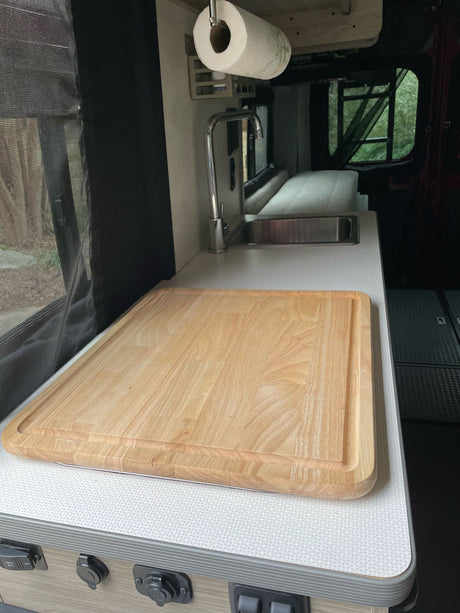 Inside an RV rental: a kitchen counter with a large wooden cutting board, sink, and screen window. Green nature outside.