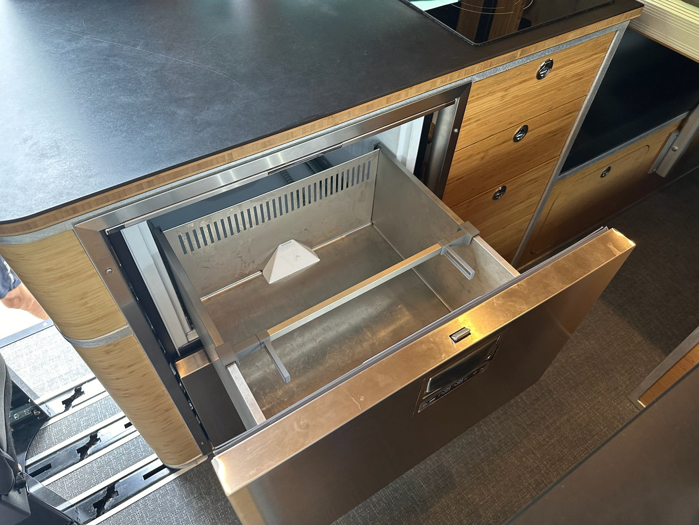View inside an RV rental's compact kitchen, featuring an open stainless steel drawer fridge, countertop, and wood storage.