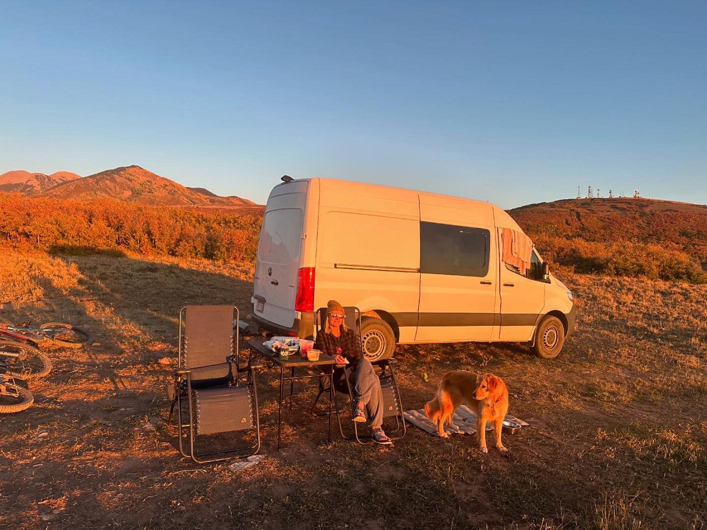 Autumn RV rental camping scene: white van, person, dog, and mountains at sunset. Perfect outdoor escape.
