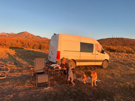 Autumn RV rental camping scene: white van, person, dog, and mountains at sunset. Perfect outdoor escape.