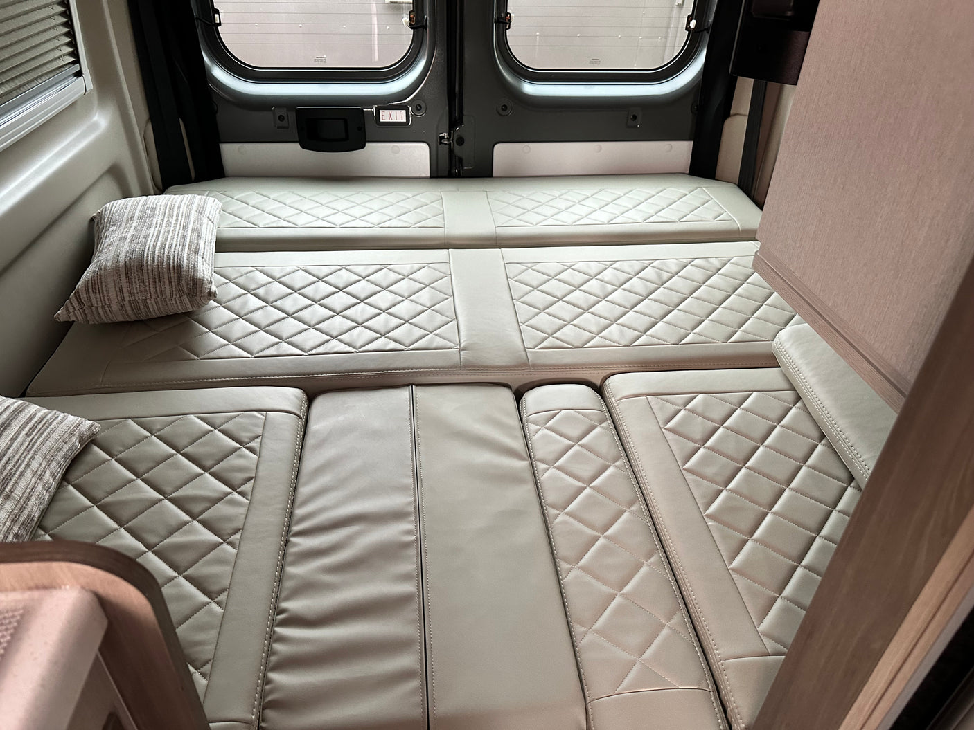 RV rental interior: Comfortable, quilted bed & seating area in a camper van, with rear windows and an "EXIT" sign.