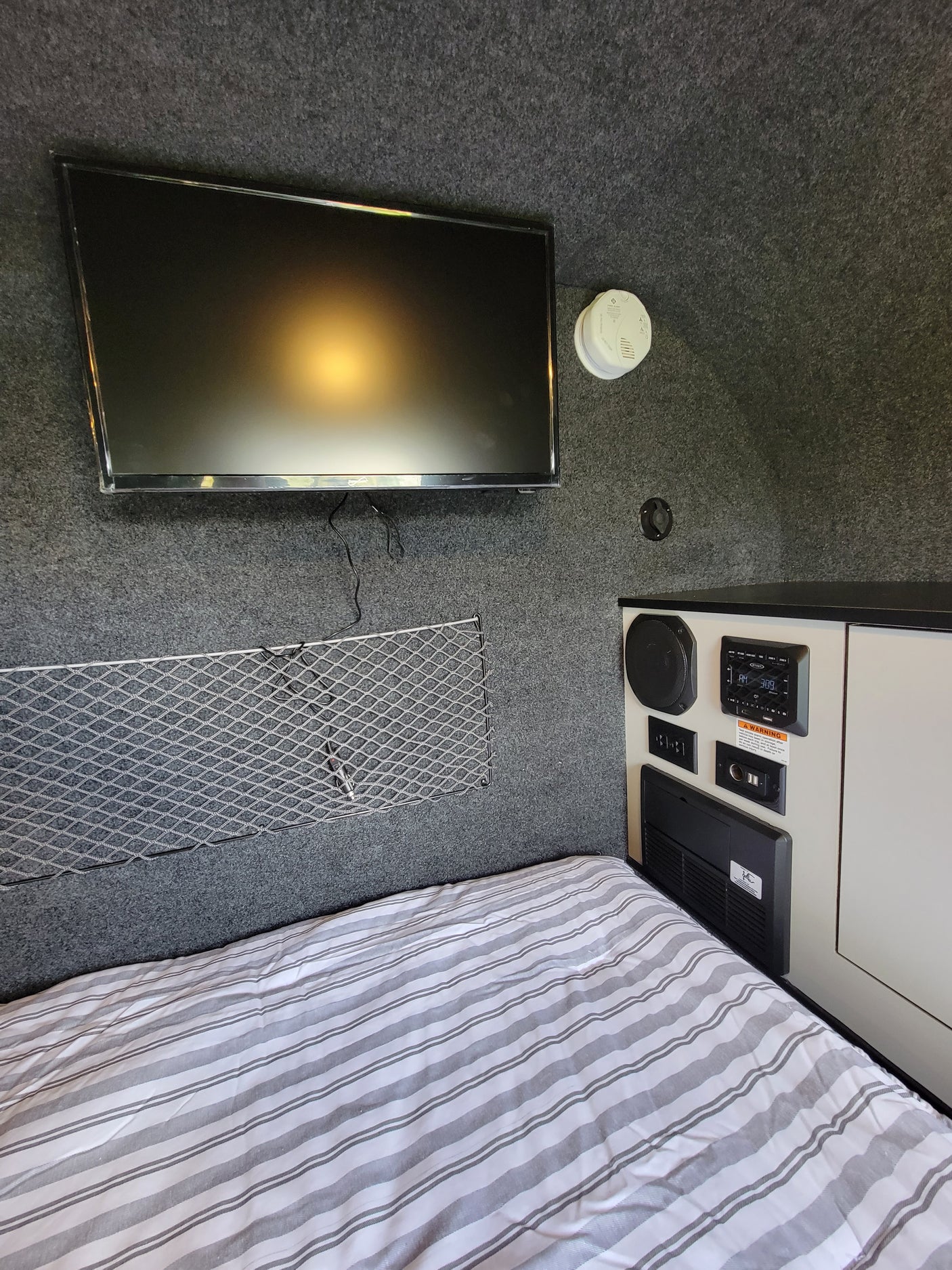 RV rental interior: TV, bed, smoke detector, and amenities control panel on gray carpet with mesh mat.