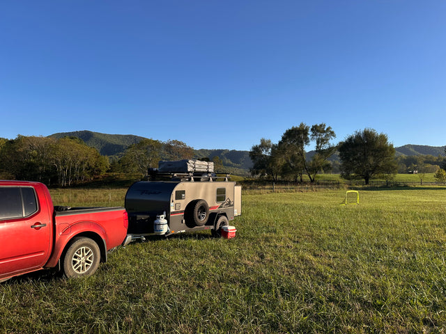 Red pickup truck and gray **RV rental** camper trailer set up in a vast green field with mountains & clear skies.