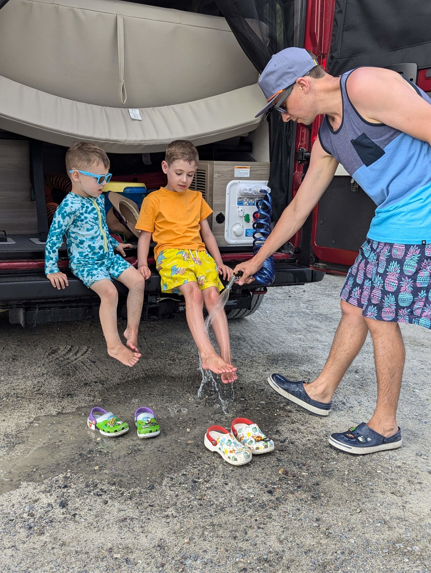 Kids get sandy feet rinsed at the back of an RV rental. A man uses an outdoor shower for the children, perfect for family adventures!