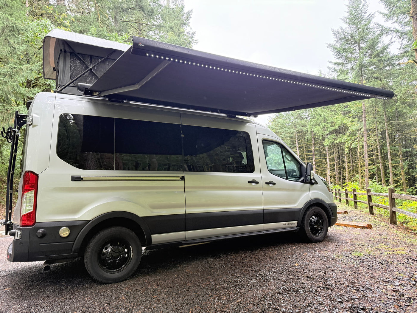 This white camper van, ideal for an rv rental adventure, features a pop-top and lighted awning in a lush forest setting. — photo 9