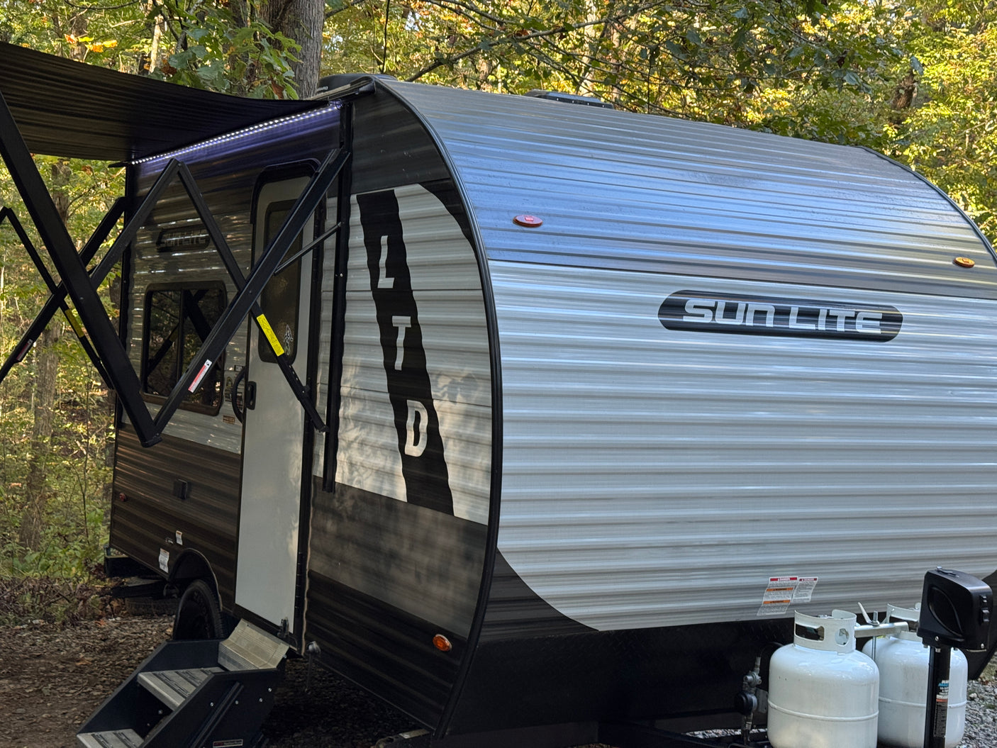 This Sun Lite travel trailer RV rental, with its awning and LED lights extended, is ready for camping in a wooded area.