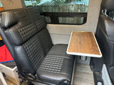 RV rental interior with a black, diamond-quilted bench seat, wooden table, and side window showing a fence.