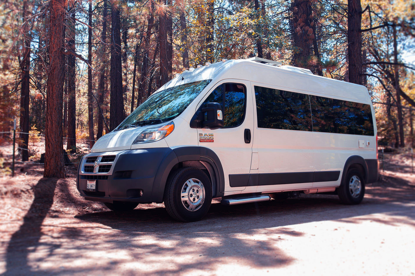 White Ram ProMaster RV rental parked on a forest trail under dappled sunlight, equipped with a roof AC and side step.