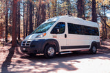 White Ram ProMaster RV rental parked on a forest trail under dappled sunlight, equipped with a roof AC and side step.