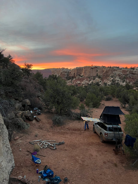 Camping under a vibrant desert sunset. An RV rental (SUV with rooftop tent) is pitched at a remote canyon campsite.