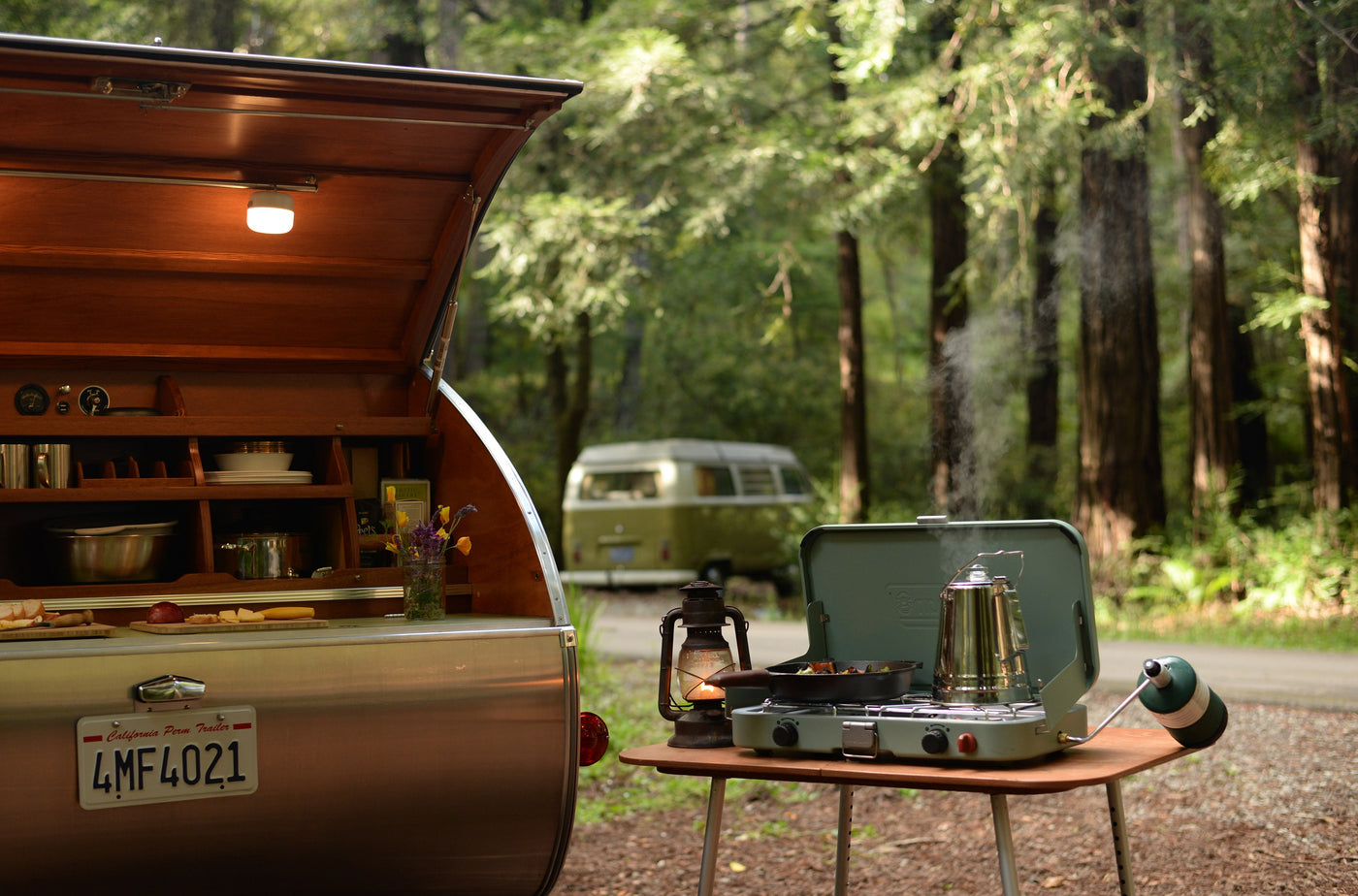 Vintage teardrop RV rental kitchen open, outdoor stove with steaming coffee, pan cooking. Forest setting with VW bus. — photo 10