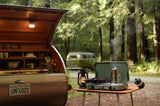 Vintage teardrop RV rental kitchen open, outdoor stove with steaming coffee, pan cooking. Forest setting with VW bus.