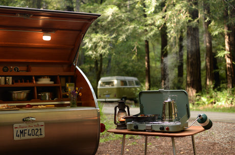 Vintage teardrop RV rental kitchen open, outdoor stove with steaming coffee, pan cooking. Forest setting with VW bus.