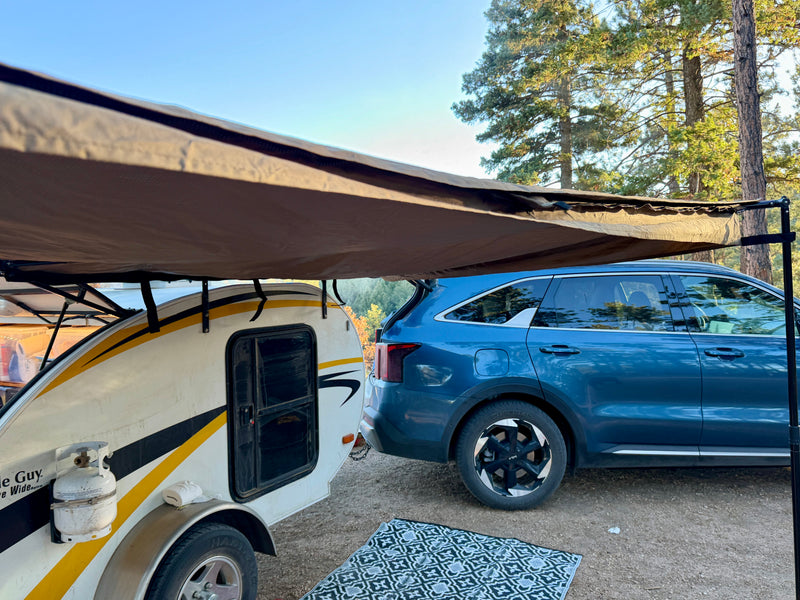 An RV rental teardrop camper with an awning extended, parked beside a blue SUV in a wooded campsite, ready for adventure. — photo 7