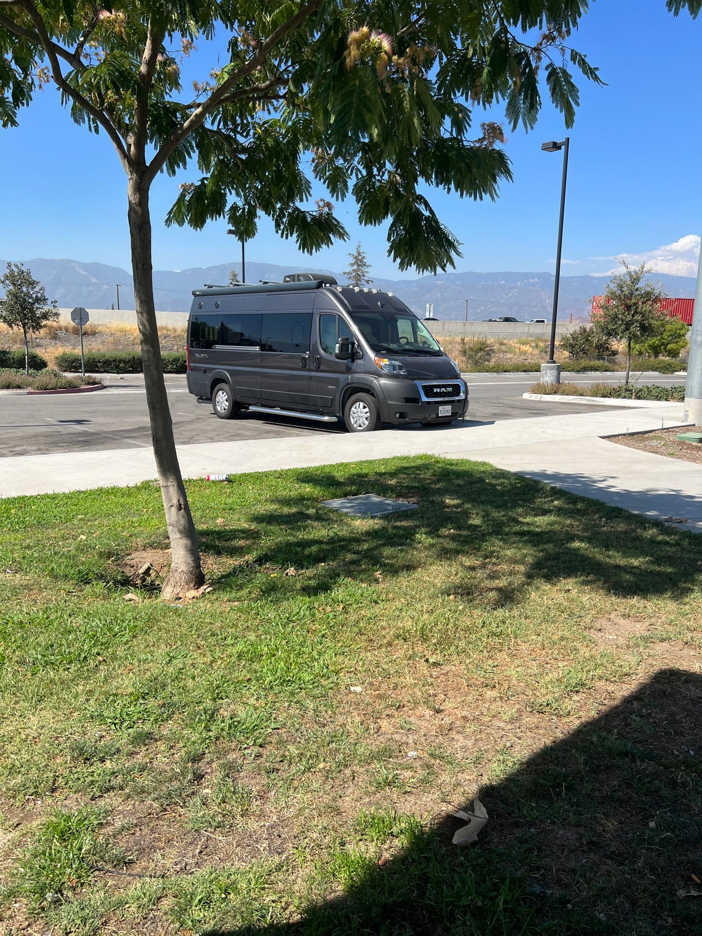 A dark grey Jayco RAM ProMaster RV rental is parked in a sunny lot, with mountains and trees in the background. — photo 8
