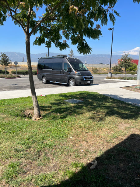 A dark grey Jayco RAM ProMaster RV rental is parked in a sunny lot, with mountains and trees in the background.