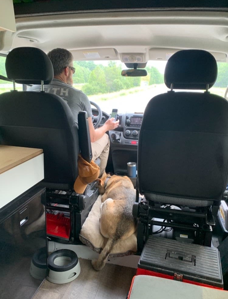 RV rental van interior: Man driving, dog asleep on cushion between front seats, dog bowls visible on floor.