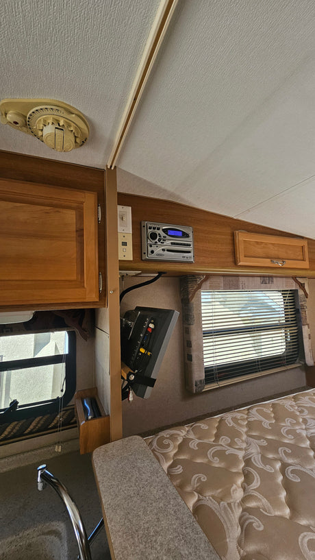 Inside a compact rv rental: wood cabinetry, Jensen entertainment system, a window, patterned bed, and kitchen area.