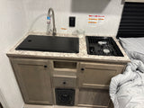 Compact RV rental kitchen with a covered sink, faucet, two-burner stove, and lower cabinets. A bed with comforter is on the right.