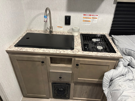 Compact RV rental kitchen with a covered sink, faucet, two-burner stove, and lower cabinets. A bed with comforter is on the right.