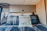 Cozy RV rental bed with "ADVENTURE AWAITS" pillow, plaid bedding, and overhead storage, ready for adventure.
