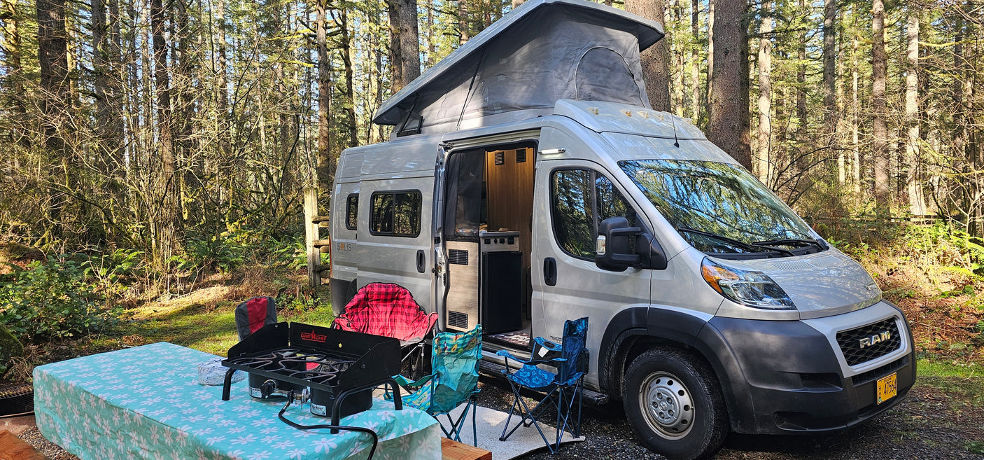 Silver RAM Solis RV rental parked at a forest campsite with a picnic table & chairs set up for outdoor cooking.
