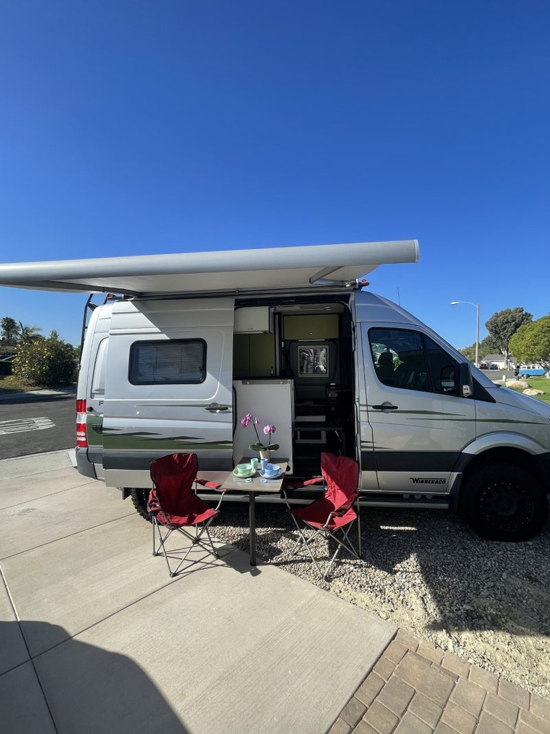 This RV rental offers a cozy outdoor setup: silver camper van, extended awning, table with flowers & red chairs. — photo 6