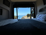 Inside an RV rental: a bed offers stunning ocean views of cliffs & blue water from open back doors.