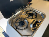 This RV rental features a compact, shiny stainless steel two-burner gas stovetop with a safety lid for cooking.