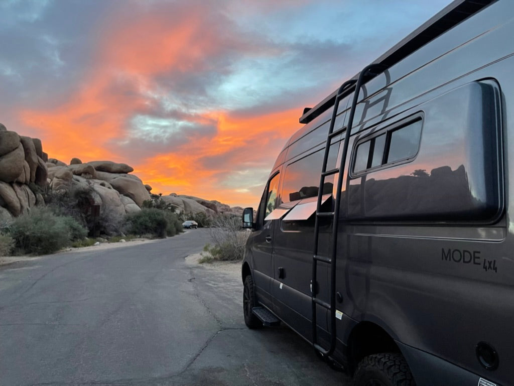 A gray RV rental van parked on a desert road at sunset. Large rock formations and vibrant orange/pink clouds fill the scene.