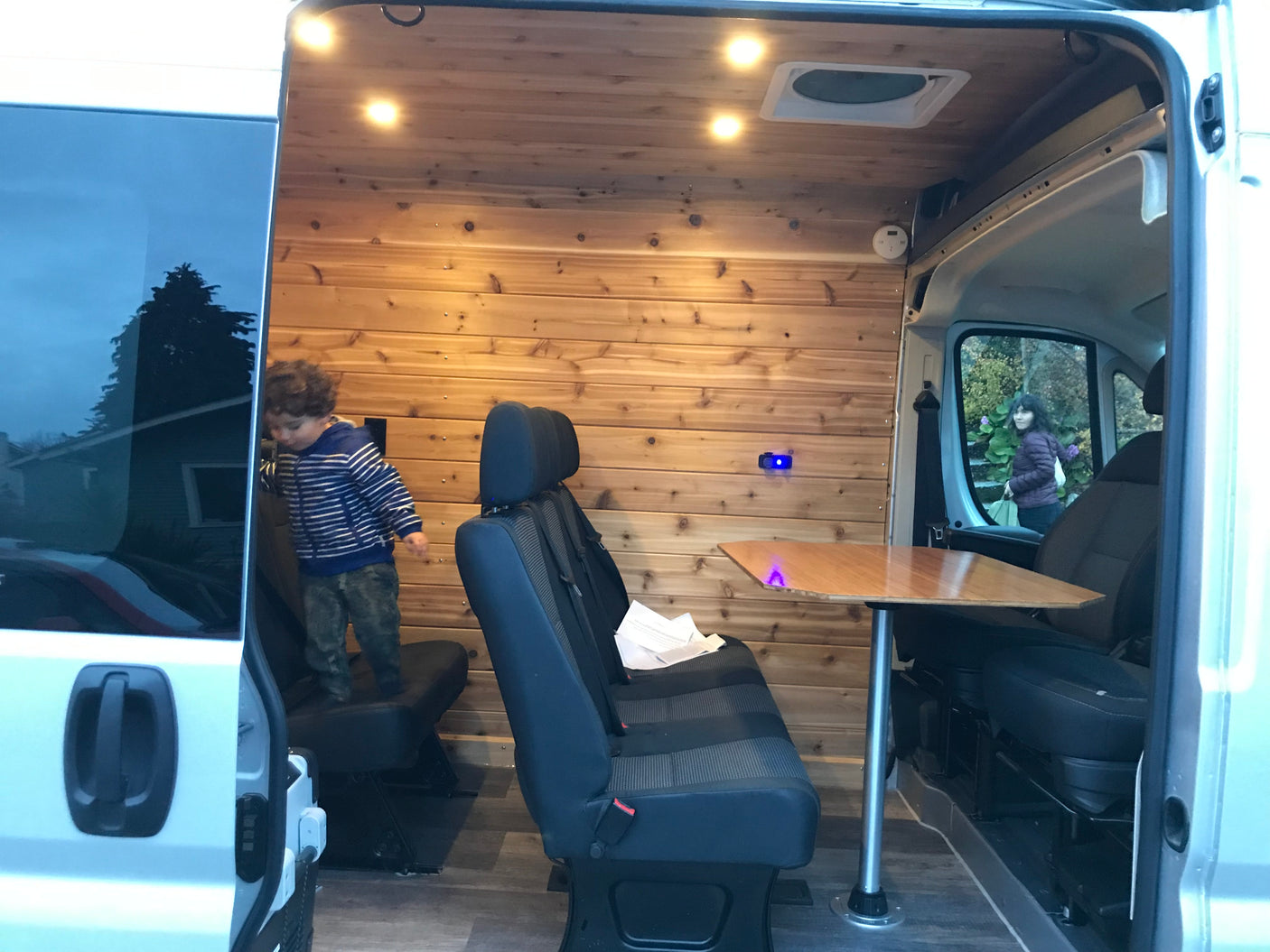 Explore this warm RV rental interior: wood walls, cozy seating, and a table. A child plays inside, a woman looks in.
