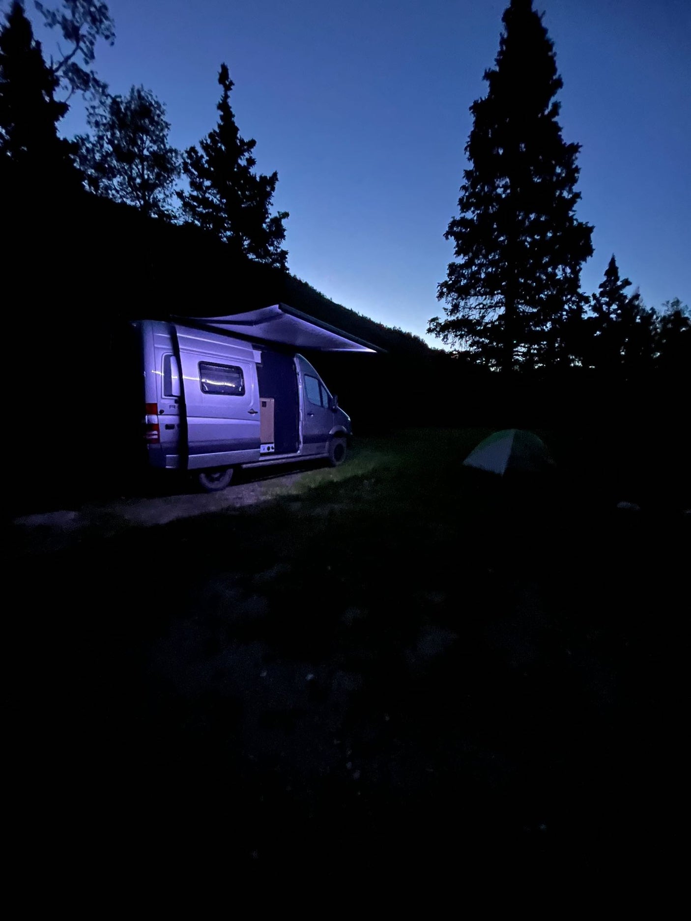 Night camping with an RV rental! A well-lit camper van with an open door and awning, alongside a small tent in a serene outdoor setting.