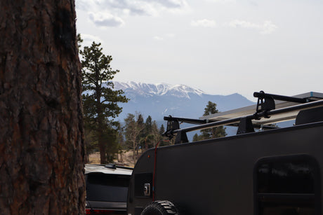 An RV rental teardrop trailer with solar panels, set against snow-capped mountains and a forest, hinting at outdoor adventures.