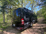 Black Airstream Interstate RV rental parked on a shaded gravel path, surrounded by green trees under a blue sky.
