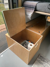Efficient RV rental storage: open wooden box shows essentials like toilet paper, next to bed with blanket. Perfect for campervan adventures.
